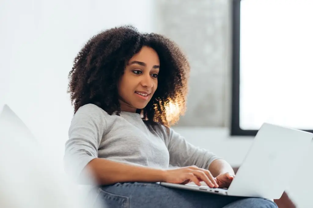 Smiling woman working remotely on a laptop, enjoying a cozy home office setup in her bright living room, embracing productivity and comfort